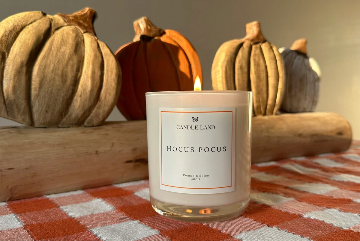 A lit Candle Land 'Hocus Pocus' pumpkin spice scented candle in a clear glass jar, set on a red and white checkered tablecloth with decorative pumpkins in the background.